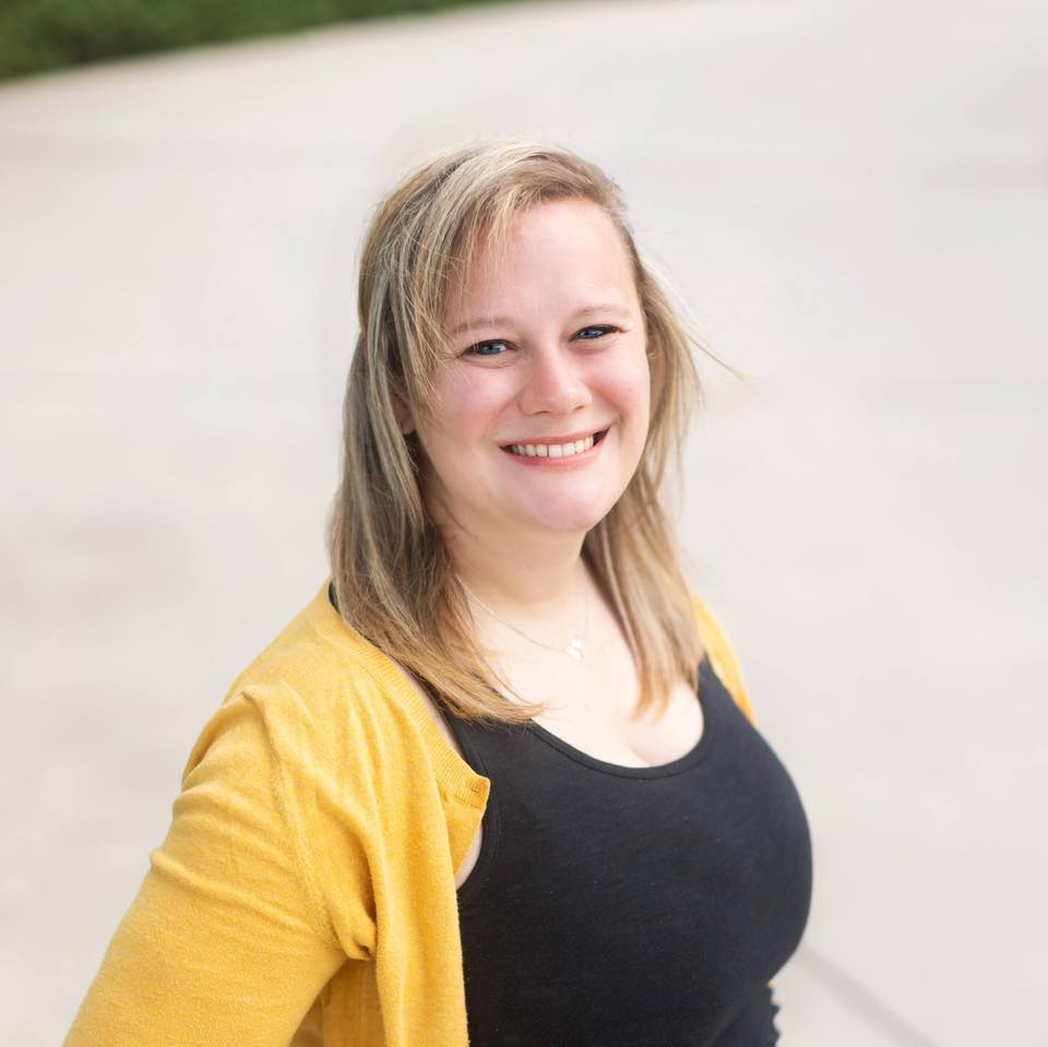 A smiling woman with blonde hair wearing a black top and a yellow cardigan stands outdoors, with a blurred background.